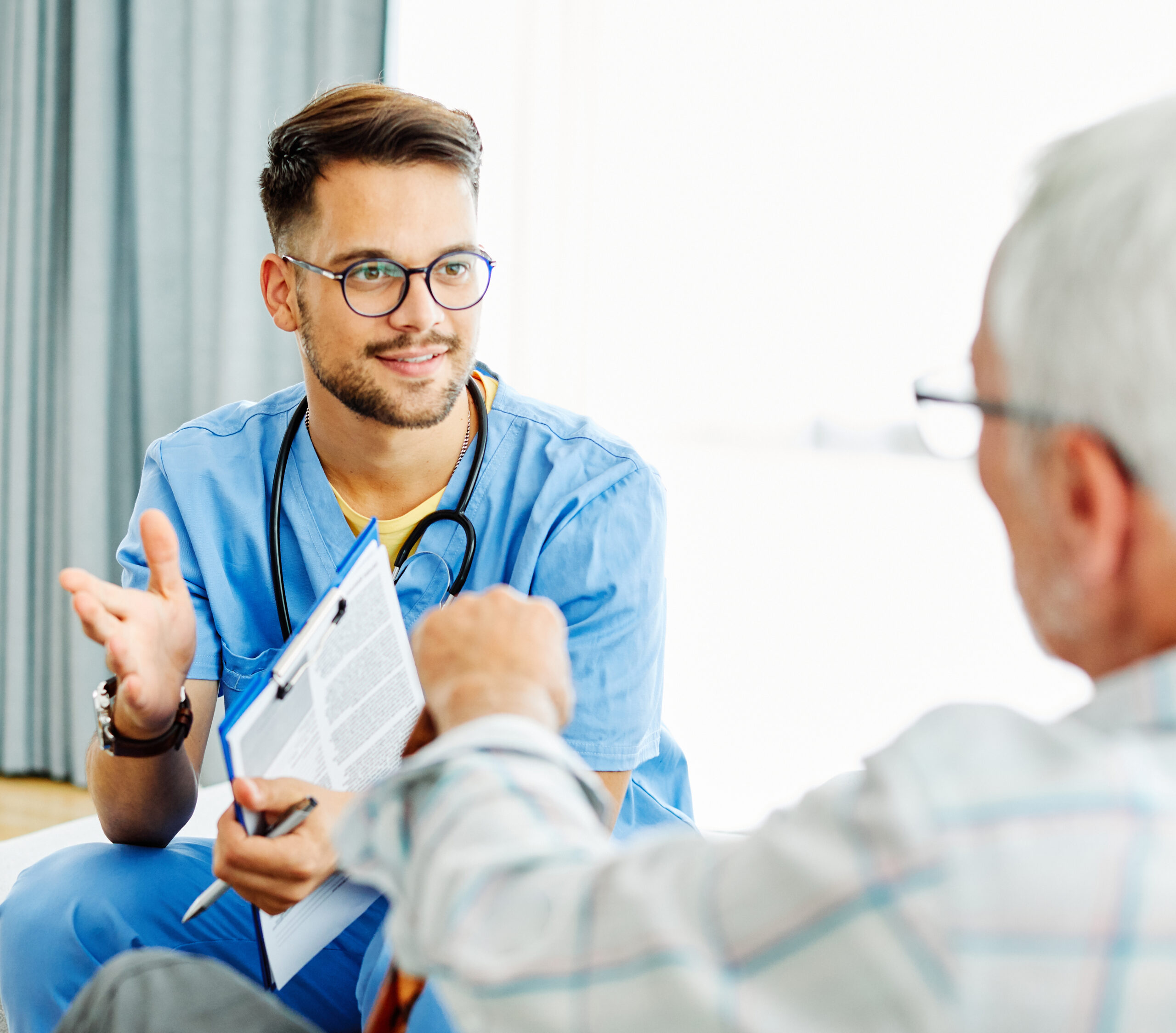 A doctor in scrubs discusses medical documents with a patient in a bright consultation room.