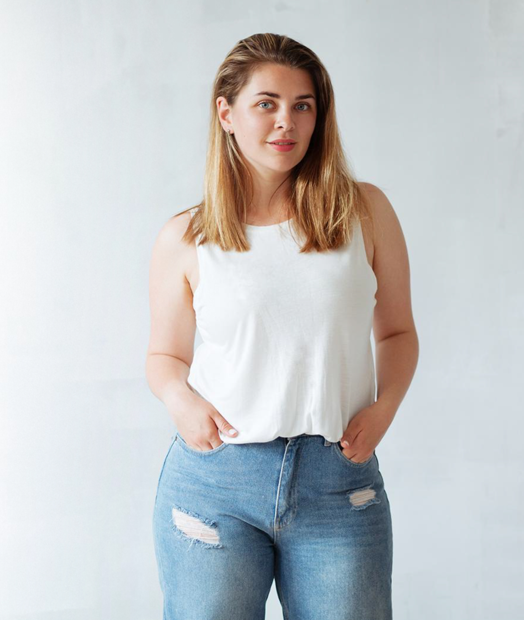 A woman posing confidently in a white top and ripped jeans against a plain background.
