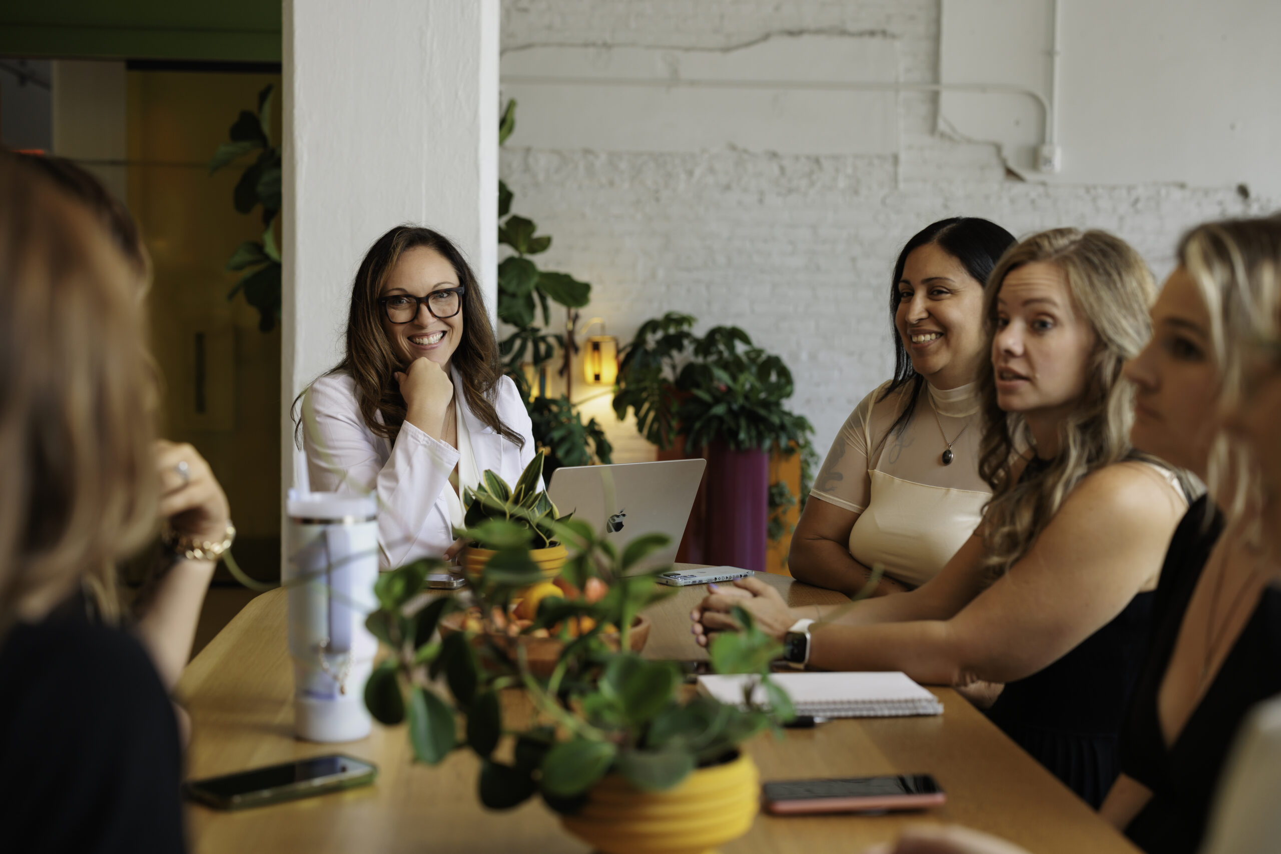 Four women engaged in a collaborative meeting around a table with plants and laptops.